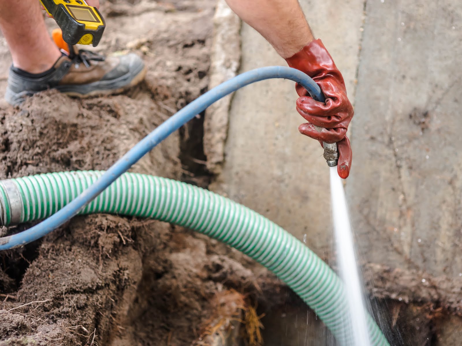 A man performing septic tank maintenance, using a hose to clean a hole in the ground.