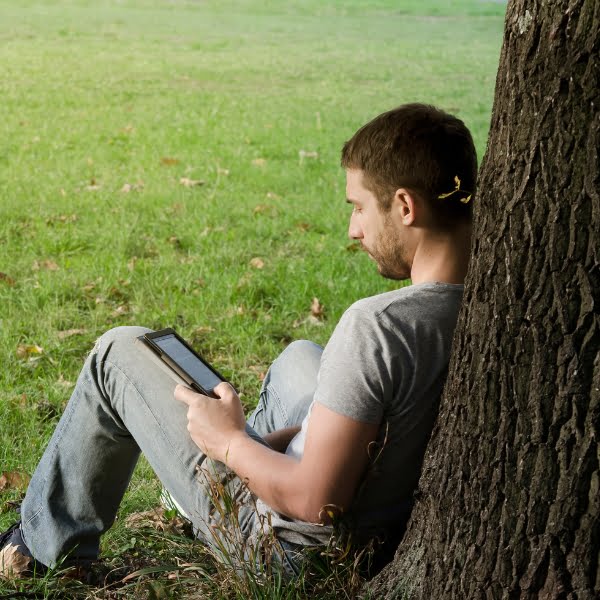 A man sitting under a tree writing a blog on his tablet computer using SEO techniques.