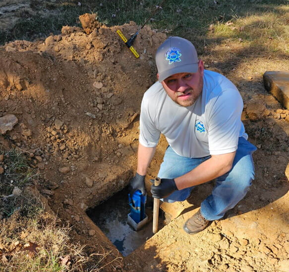 A septic tank company employee kneeling in a hole with a shovel.
