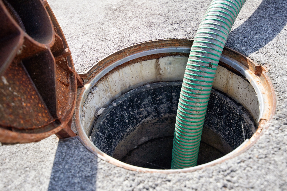 A flexible green hose extends into a manhole with rusted edges on an asphalt surface for emergency septic services.