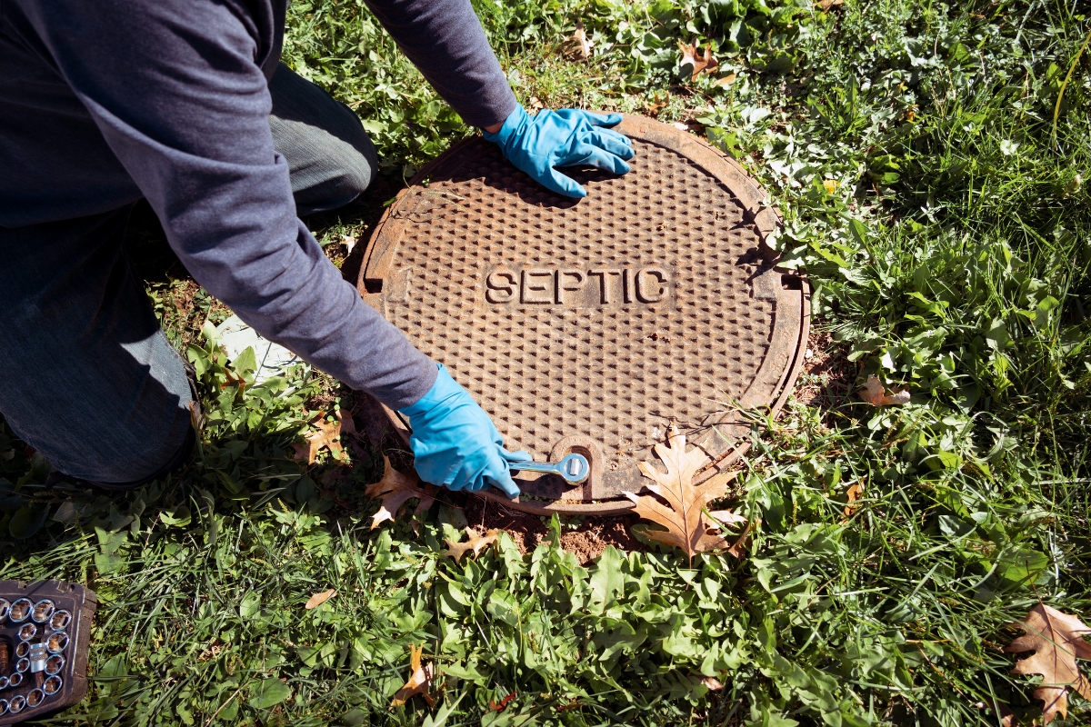 A person wearing gloves adjusts a septic tank cover in a grassy area, with tools and materials for the septic system installation nearby.