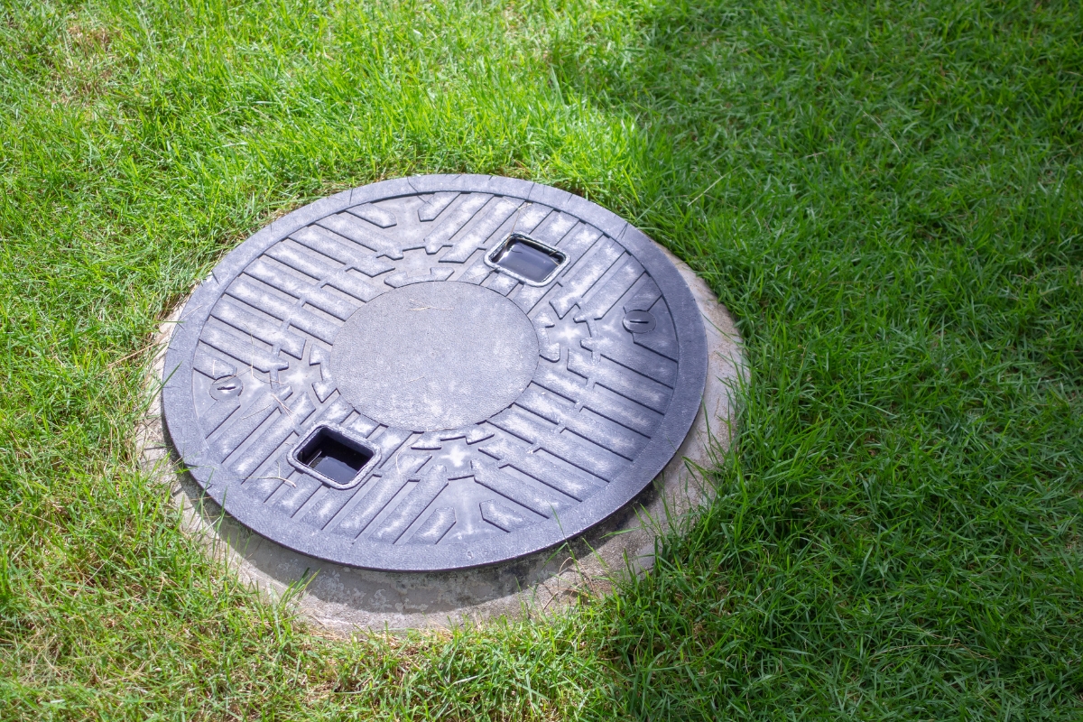 A round, metal commercial septic system cover embedded in a grassy lawn under daylight.