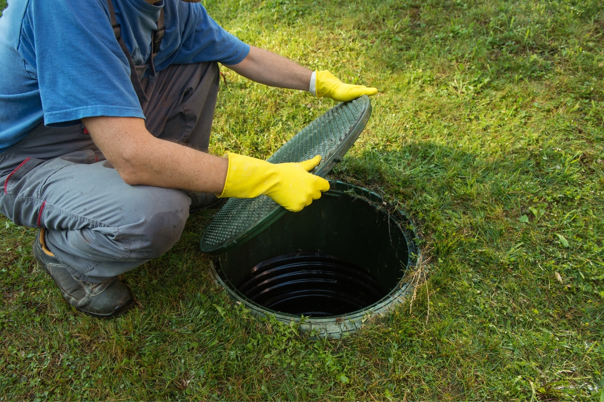A worker in blue attire and yellow gloves opens a round, green commercial septic system cover on a grassy area.