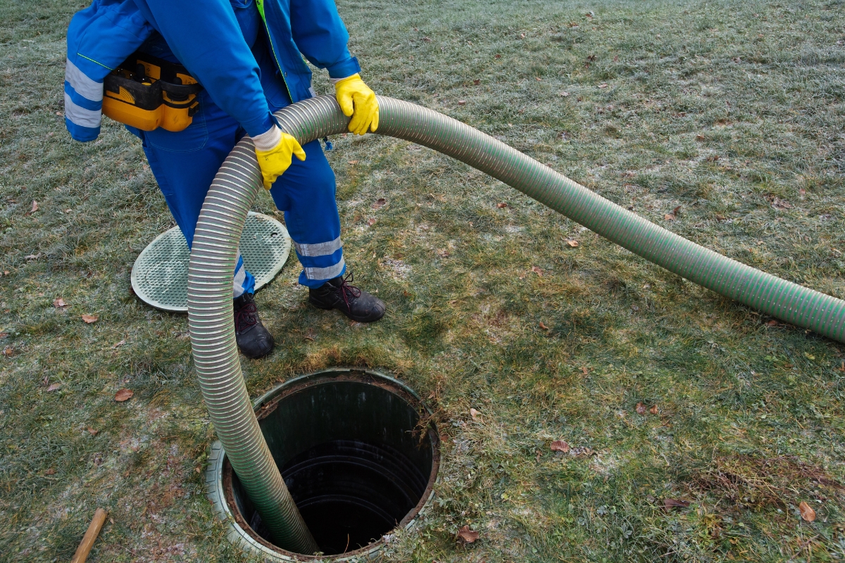 Worker in blue coveralls and yellow gloves using a large hose to perform maintenance on commercial septic systems through an open manhole.