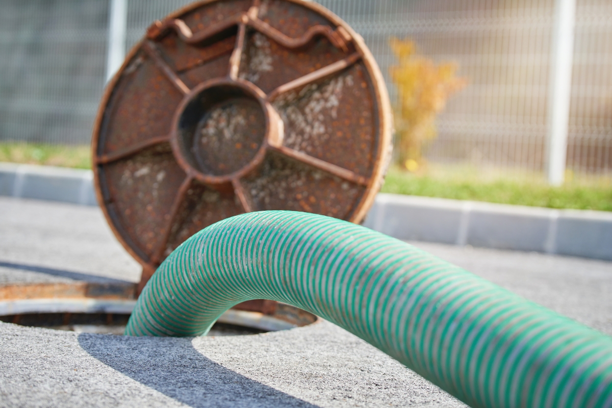 A green industrial hose extends into a rusty round metal hatch on a paved surface, illustrating maintenance of commercial septic systems.