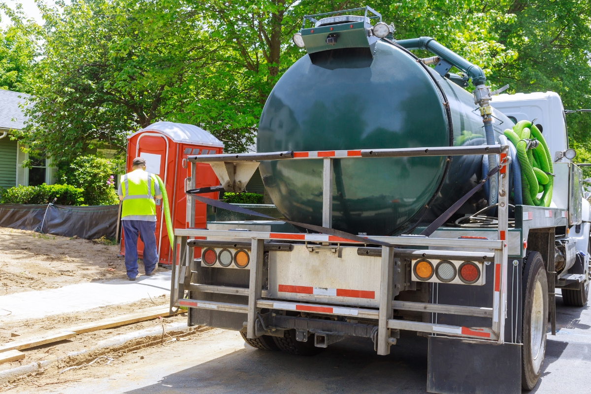 A worker in a high-visibility vest walks near a commercial septic systems truck and a portable toilet beside a residential street under construction.