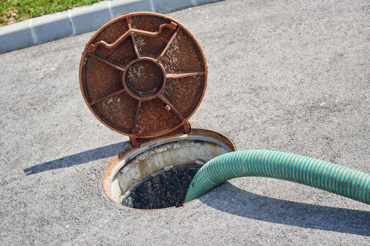 A green hose running into an open, rusted round manhole cover on a concrete surface, part of a commercial septic system.