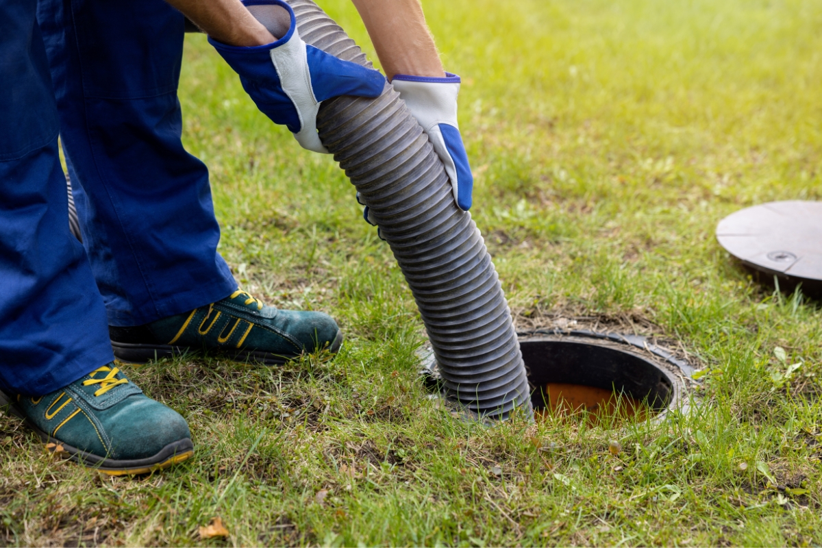 Worker in blue coveralls and safety shoes uses a large flexible hose to inspect or clean a commercial septic system opening in a grassy area.