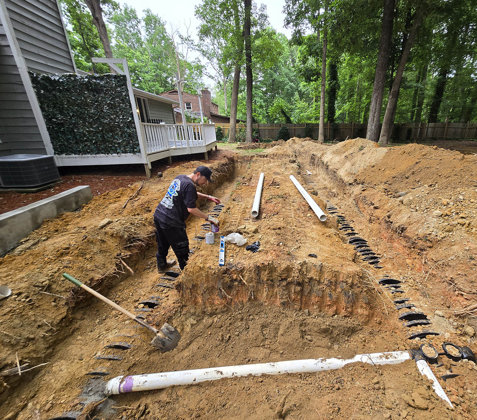 A person works in a deep excavated site next to a house, surrounded by trees. Various pipes and tools from a septic tank company are scattered around the area.