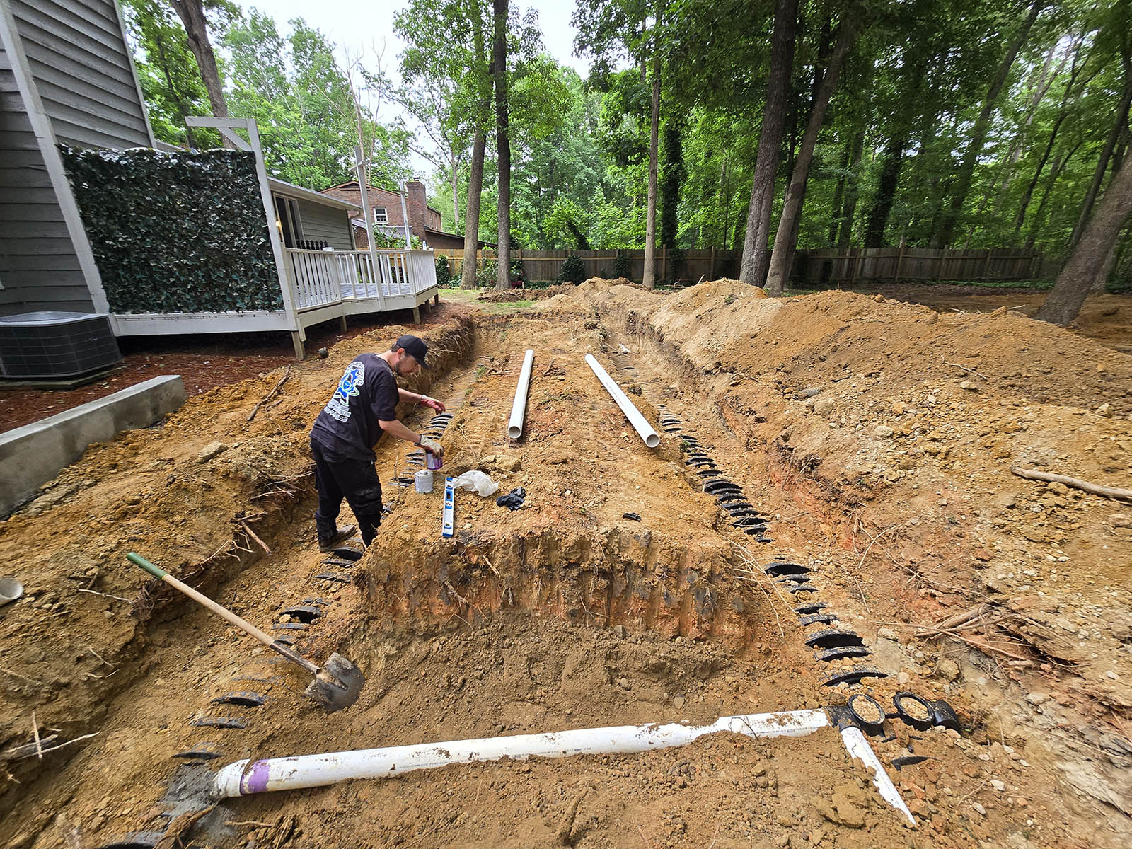 A person is working in a trench-filled yard with visible drainage pipes, presumably for a septic tank company. A house with a white porch stands on the left, surrounded by trees.