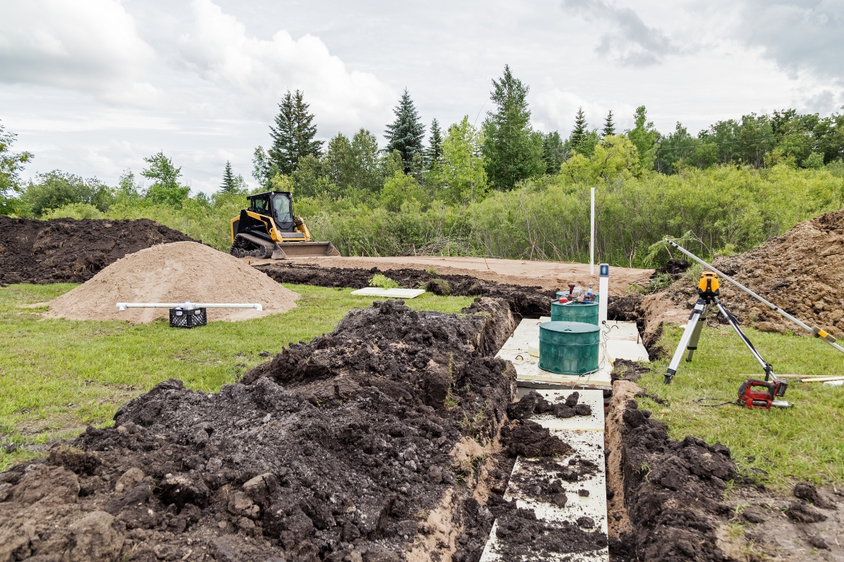 Construction site with a bulldozer, excavated soil, exposed piping for water conservation, and surveying equipment in a grassy field.