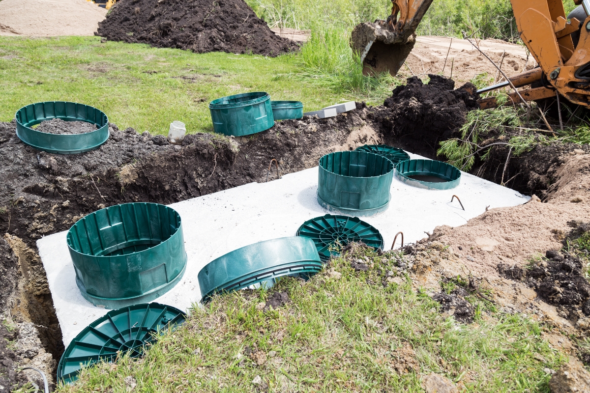Green plastic water conservation modules on a white geotextile fabric next to a trench being dug by a backhoe in a grassy area.