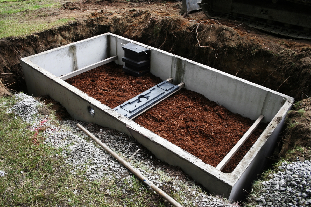 An open rectangular concrete vault partially buried next to an excavator, filled with water conservation equipment and reddish-orange organic material, with various construction tools on the ground.