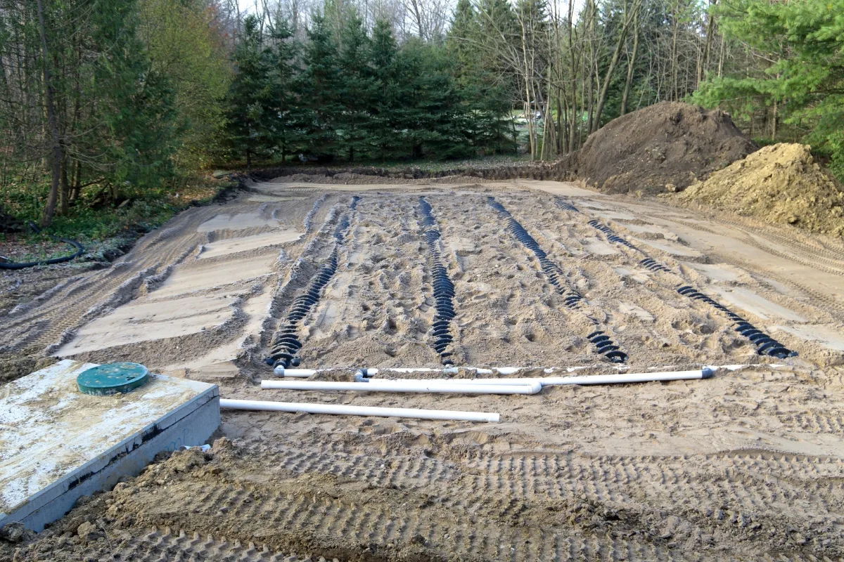 Water Conservation with Septic Systems: A Green Approach to Wastewater 1 A construction site with tire tracks on sandy soil, surrounded by trees, featuring exposed pipes and a pile of dirt, emphasizing water conservation efforts.