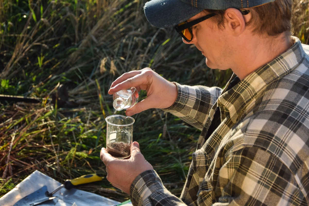 Person wearing a flannel shirt, cap, and sunglasses pours liquid from a small container into a beaker with soil, likely conducting an outdoor experiment or soil testing for septic systems. Tools are nearby on a mat.