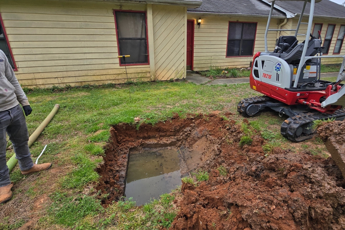 A backyard with a large, water-filled hole being excavated next to a yellow house. A mini excavator is parked nearby, and a worker stands at the edge of the hole, preparing for an eco-friendly green septic system installation.