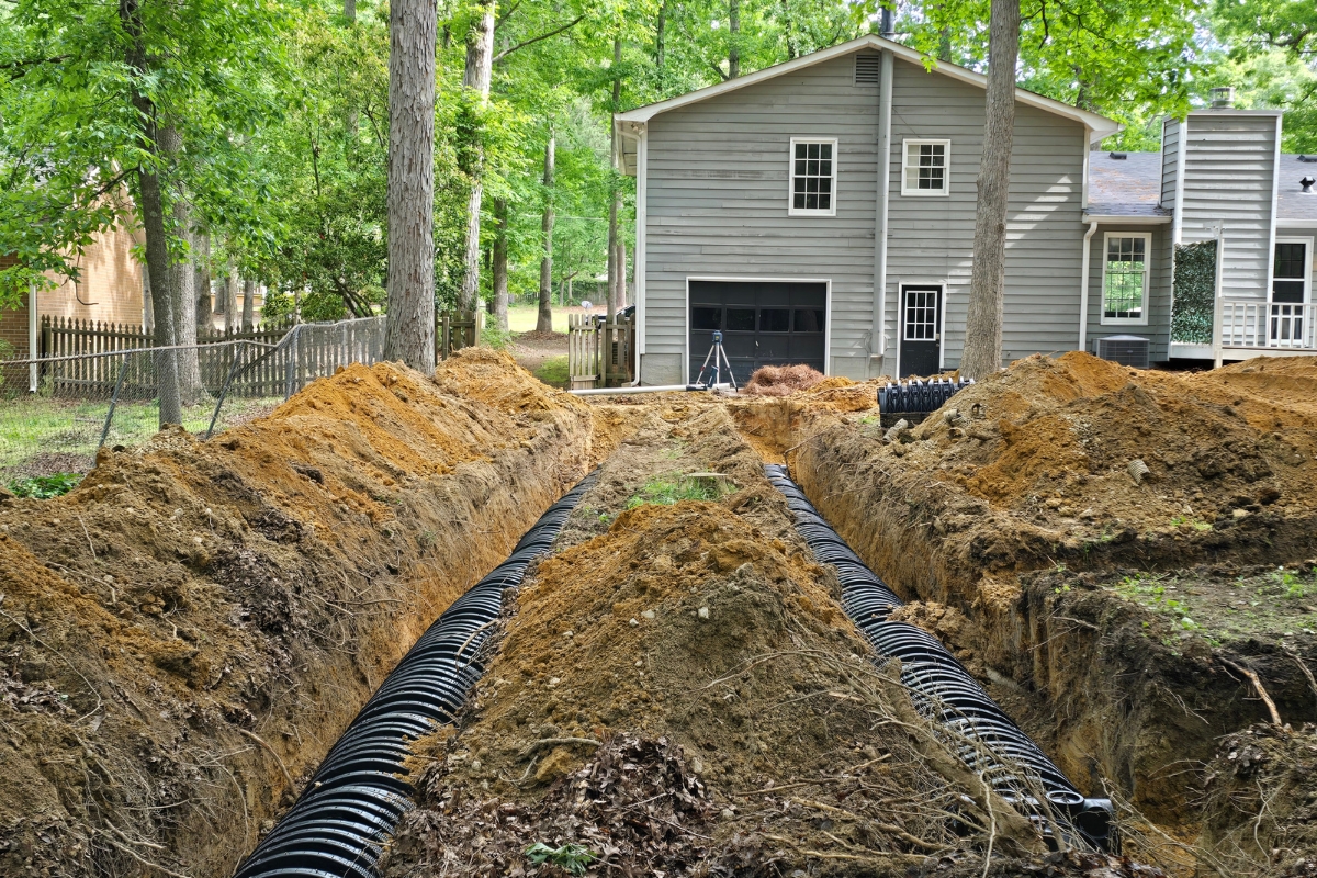 A grey house with construction trenches and drain pipes being installed in its backyard, among trees, is also incorporating green septic systems for an eco-friendly touch.