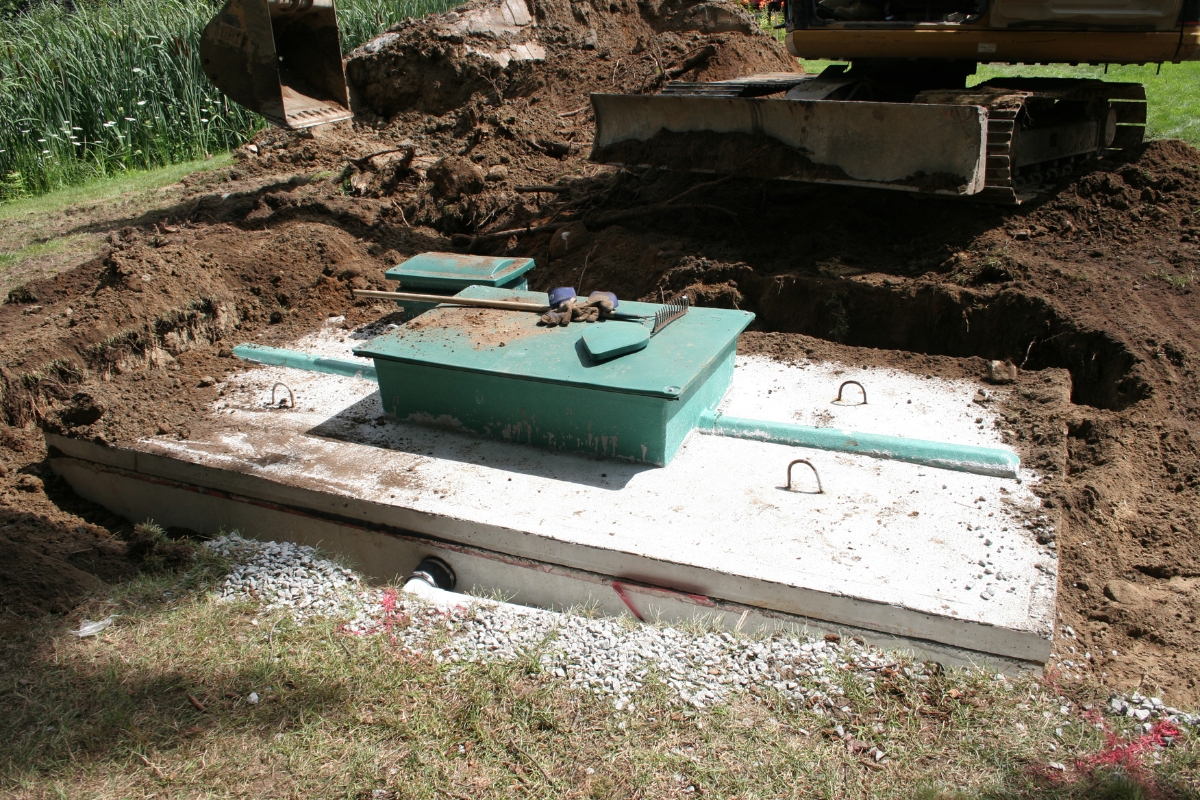 A partially installed septic tank is visible, surrounded by dirt and a construction vehicle. The septic tank lid is open, and various tools are placed on and around it. This setup highlights the initial steps in implementing green septic systems for sustainable waste management.