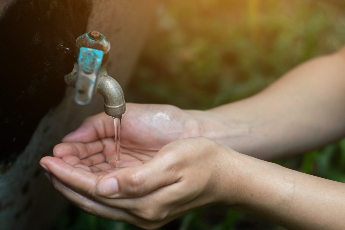 A person's hands are cupped under a small metal faucet with water trickling out, set against a blurred outdoor background, highlighting the importance of green septic systems for sustainable living.