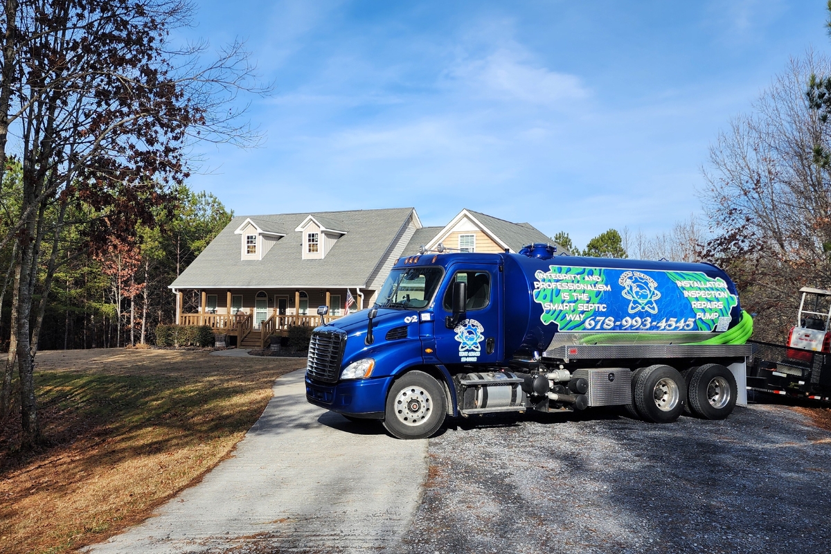 A blue septic service truck with business contact information is parked on a gravel driveway in front of a large, renovated house with a porch on a clear day.