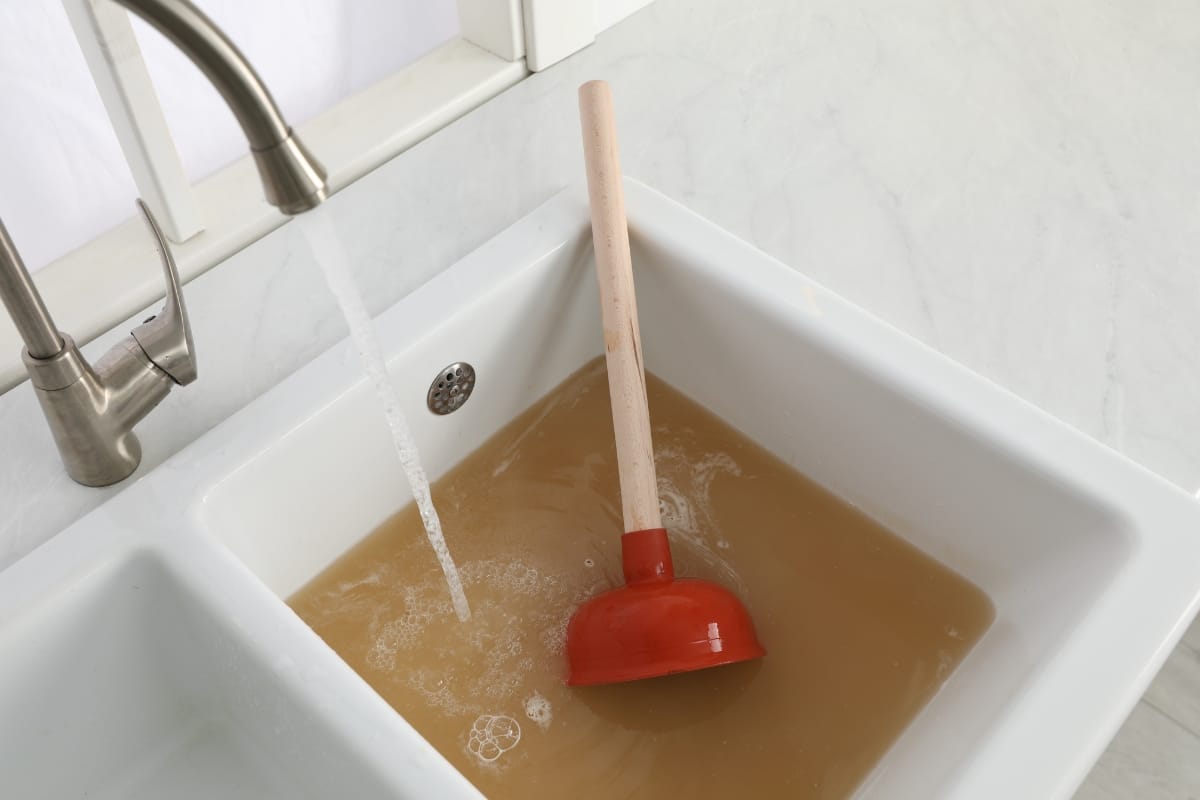 A kitchen sink filled with dirty brown water and a plunger submerged in it, with the faucet running, hints at ongoing home renovation challenges. It's a scene that may even make you question the state of your septic system.