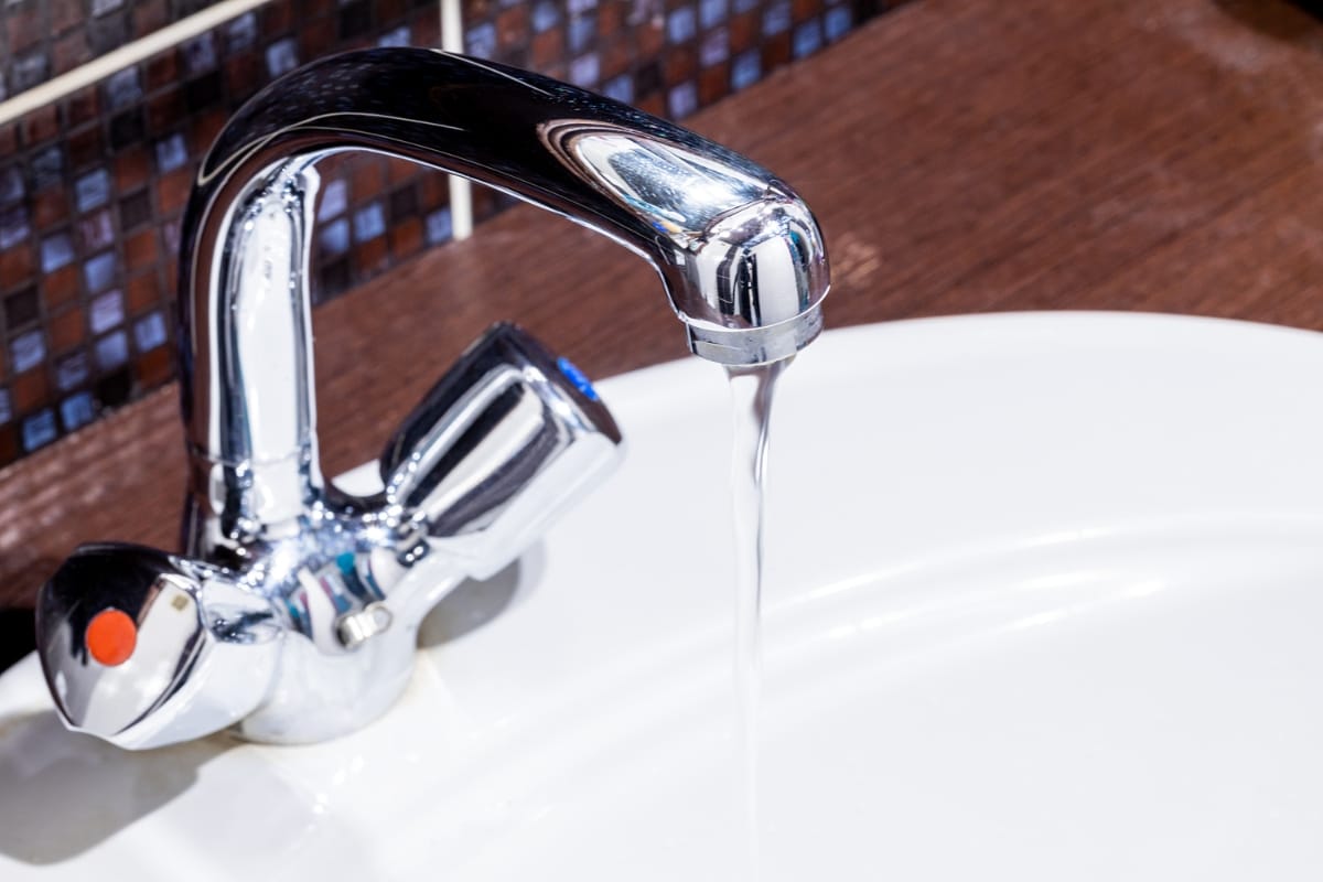 A chrome faucet with a single red dot lever dispenses water into a white sink, seamlessly integrating into your home renovation. Dark mosaic tiles serve as the perfect backdrop.