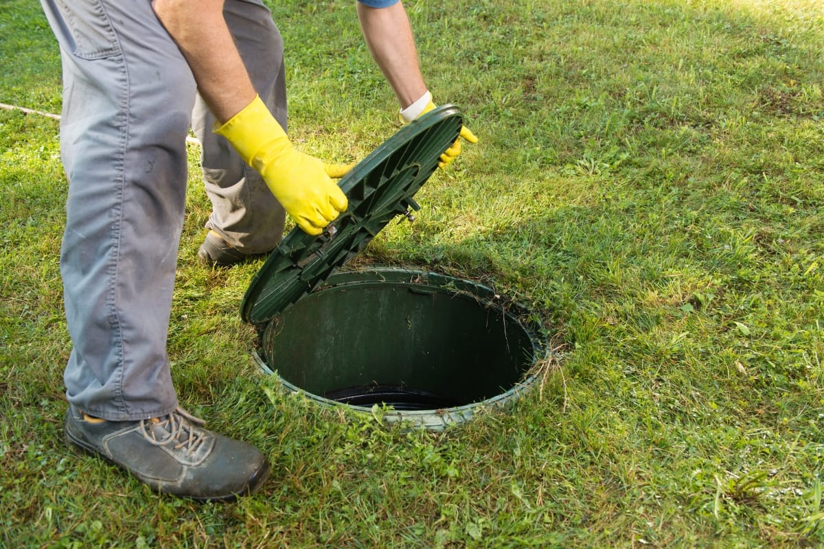 A person wearing yellow gloves and gray pants lifts the lid off a green outdoor septic tank on a grassy lawn, likely part of an ongoing home renovation project.