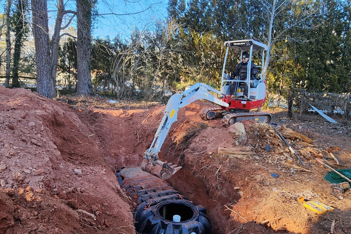 A person operates a small excavator to cover a partially buried septic tank in a red dirt area surrounded by trees, as part of an ongoing home renovation.