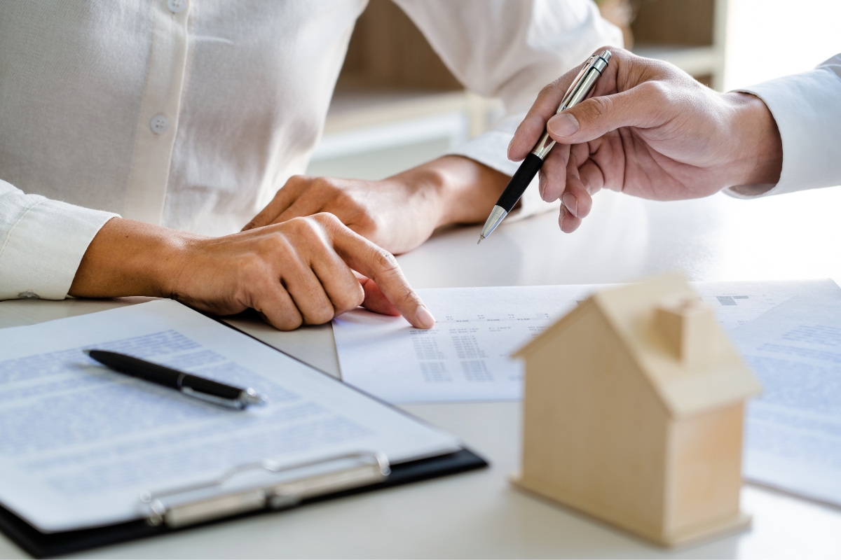 Two people reviewing real estate documents at a desk with a miniature wooden house in the foreground. One person points at the paper while the other holds a pen. A clipboard and pen are also on the desk, possibly discussing important aspects of septic systems.