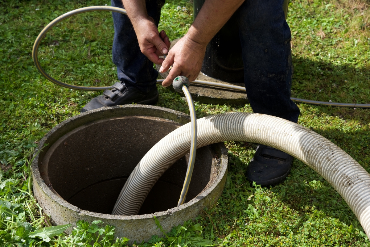 A person is carefully inserting a hose into a circular underground opening, possibly a septic tank, on a grassy lawn, demonstrating the importance of maintaining septic systems in real estate.