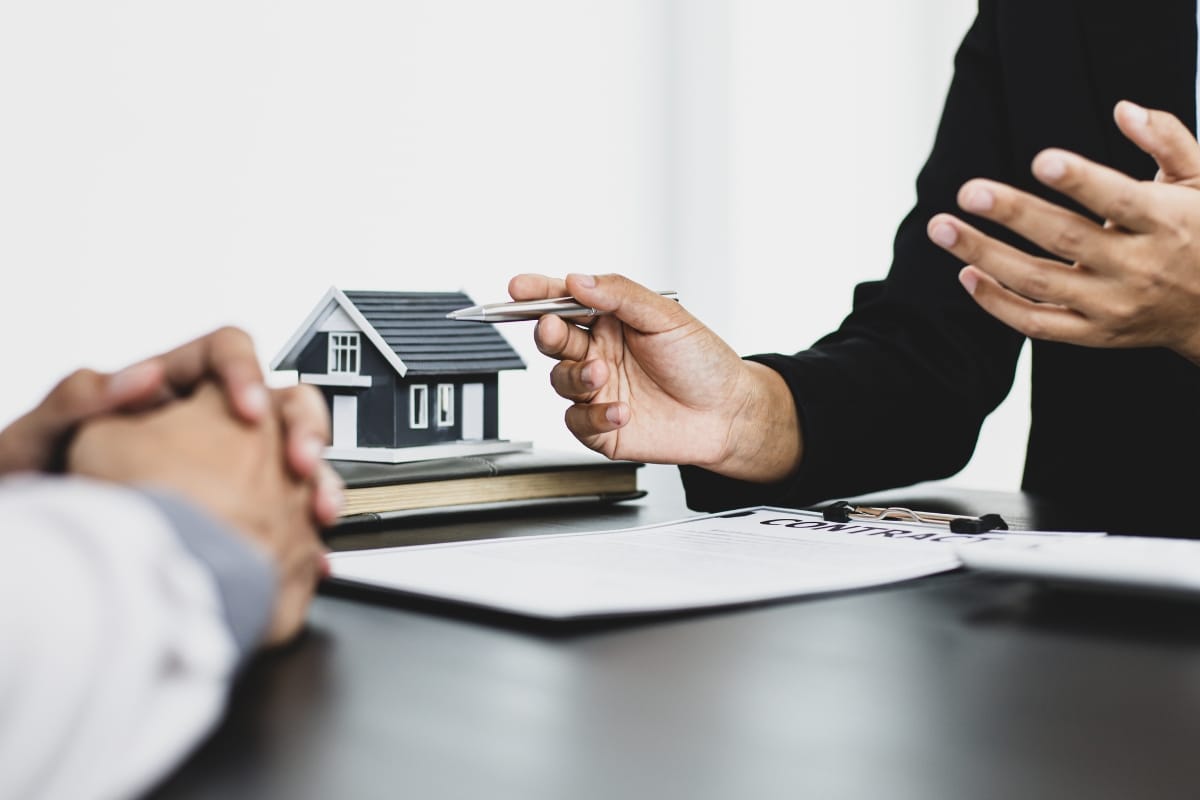 Two people are seated at a table discussing real estate, with a model house and documents in view. One person is holding a pen, gesturing during the conversation about the importance of septic systems in their property decisions.