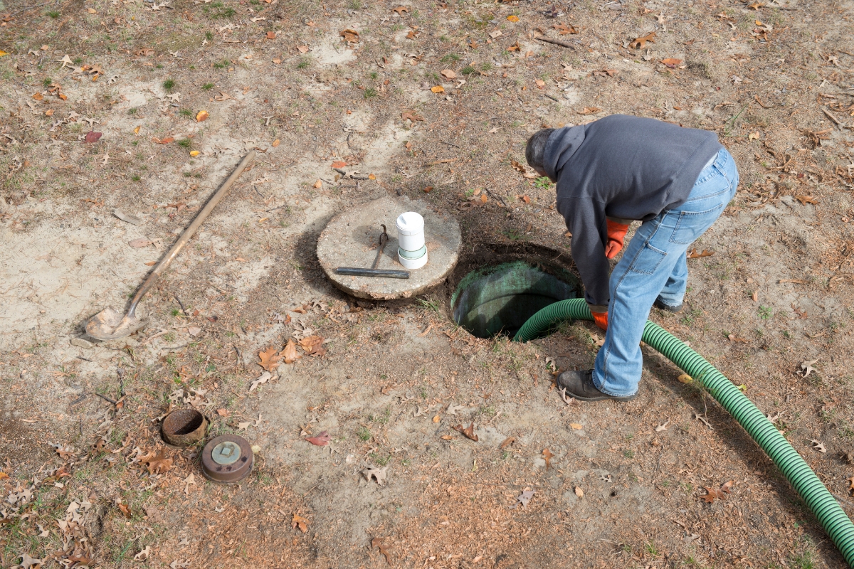 A person in a hoodie and jeans works on a rural septic system, using a large green hose, with a shovel and tools nearby on dry, patchy ground.