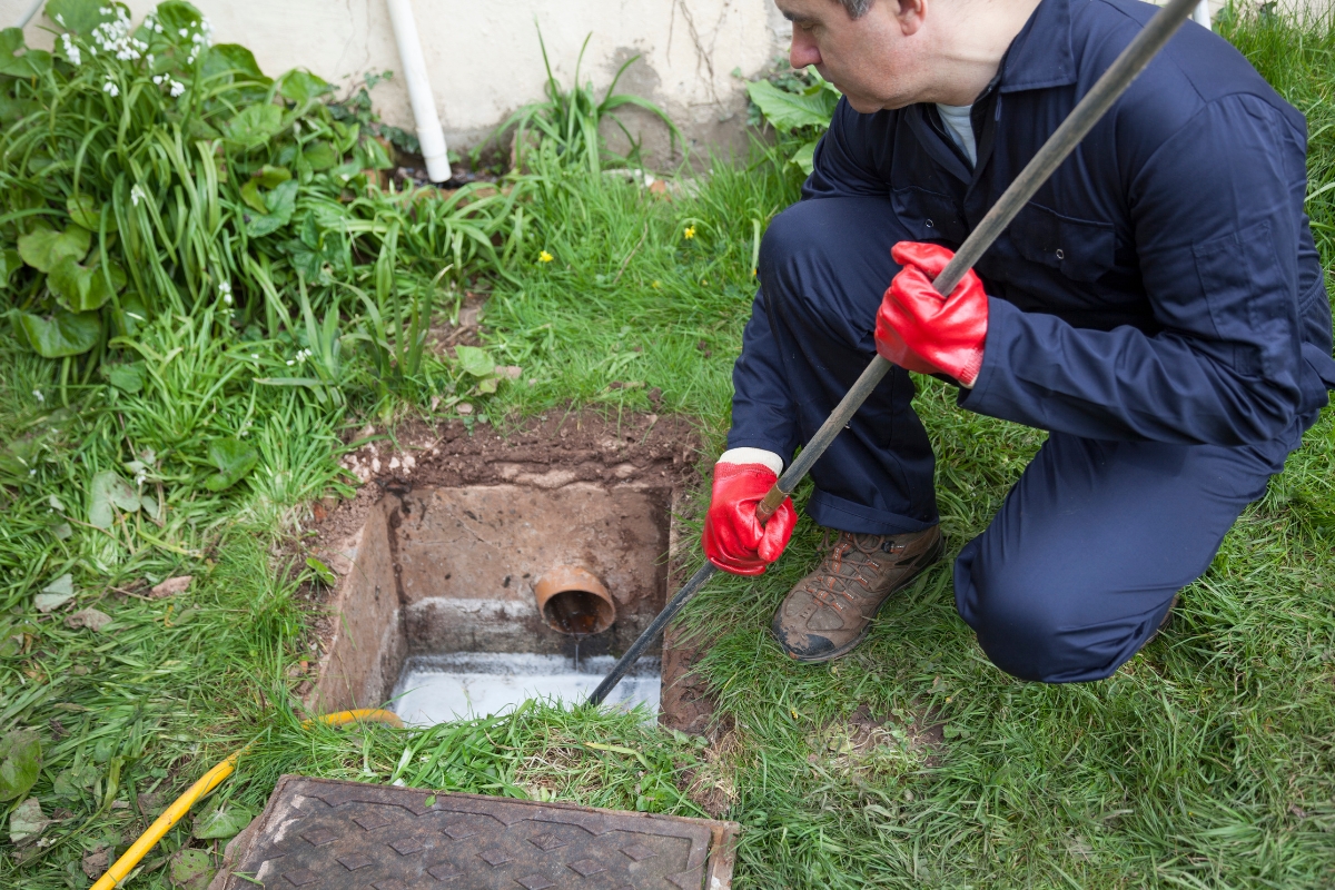 A person wearing a blue jumpsuit and red gloves uses a long rod to inspect or clean a drain in a grassy area, likely maintaining rural septic systems.