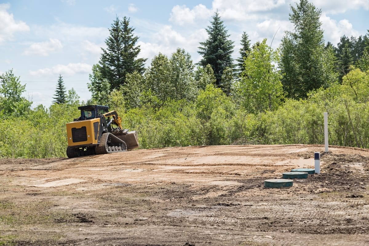 A yellow bulldozer spreads dirt on an excavated area for rural septic systems, surrounded by green trees and bushes on a sunny day.