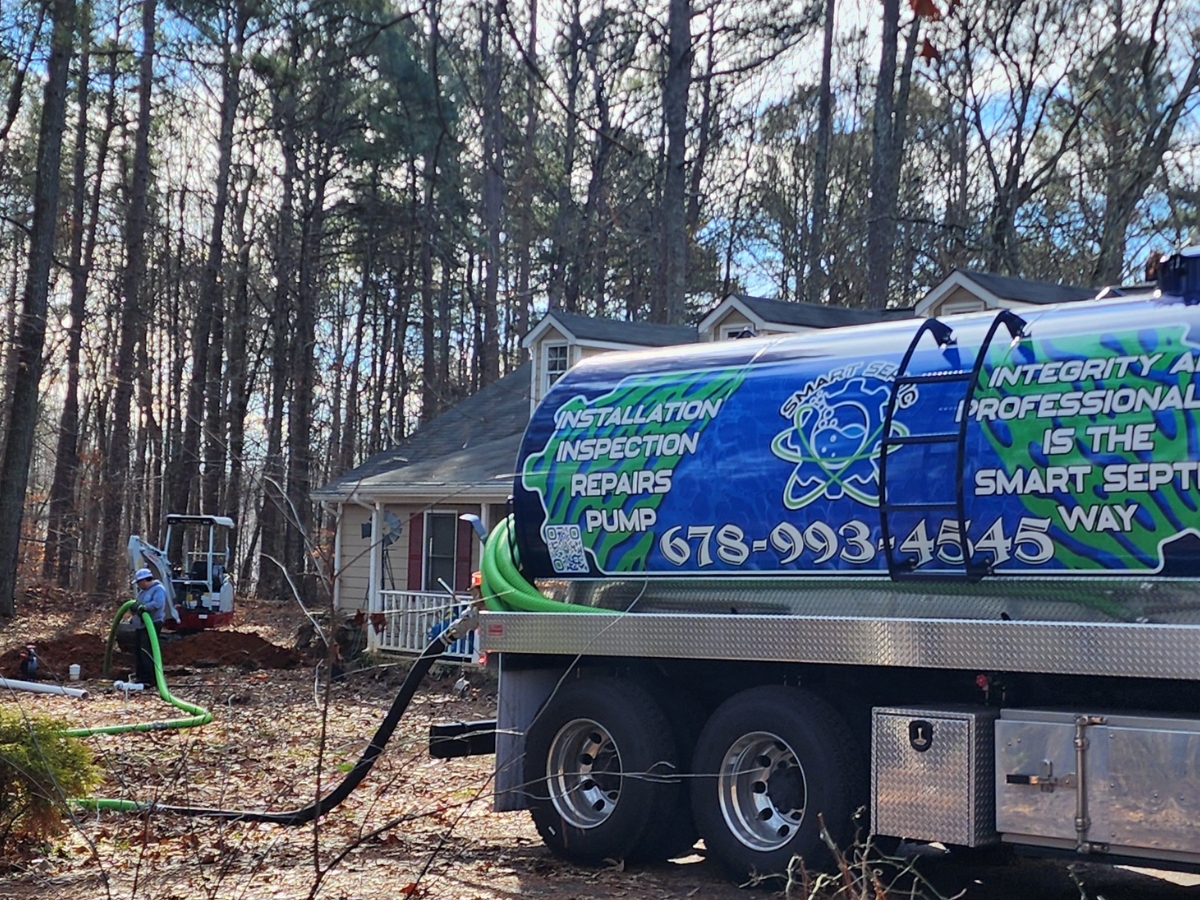 A septic service truck is parked in front of a house surrounded by trees, with hoses connected to the septic system. A small excavator is in use nearby, demonstrating their commitment to 24-hour septic service.