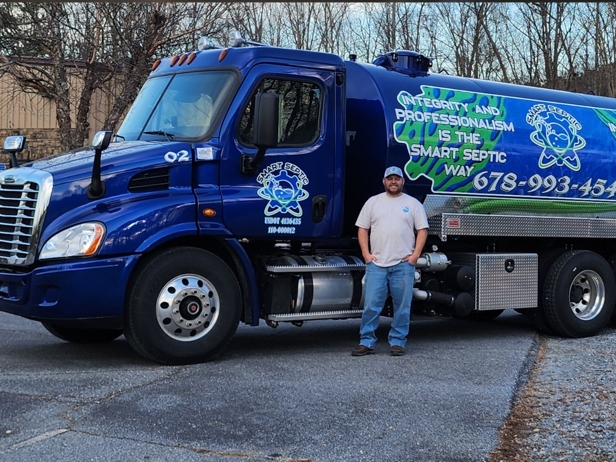 A Smart Septic Pro expert in a light-colored shirt and cap stands beside a blue septic service truck adorned with company logos, emphasizing their expertise in septic tank planning and septic system repair. Trees and a building can be seen in the background.