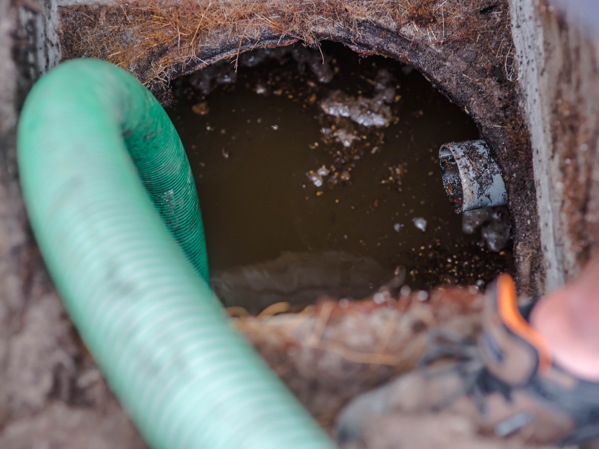 A close-up view of a green hose inserted into a septic tank for emergency septic tank cleaning, with murky water and dirt visible inside the tank. Part of an individual's shoe is seen at the bottom right corner.