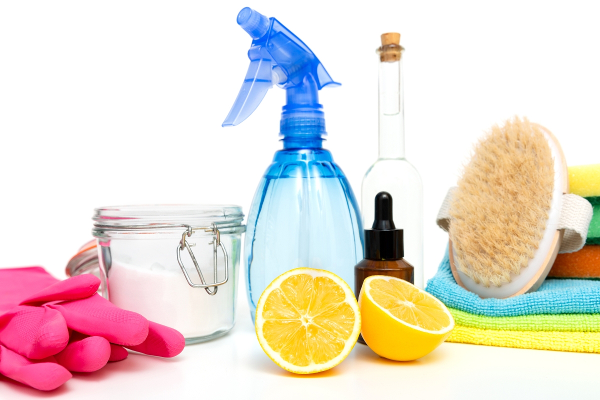 Assorted cleaning supplies ideal for everyday and septic tank cleaning, featuring a spray bottle, lemon halves, jar of powder, glass bottle, dropper bottle, scrub brush, gloves, and folded towels arranged on a white background.