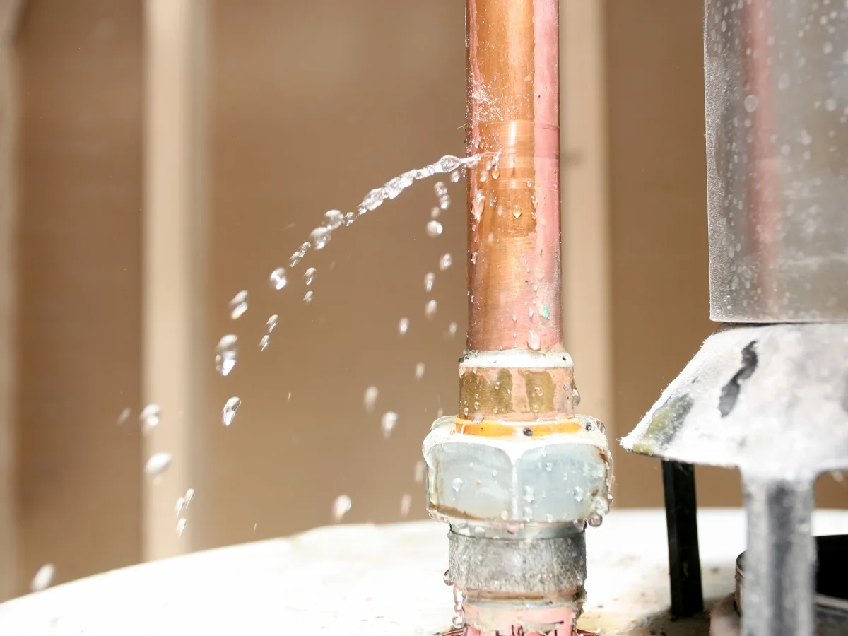 Water is spraying out from a small leak in a copper pipe connection inside the building, necessitating an emergency septic tank cleaning to prevent any further complications.