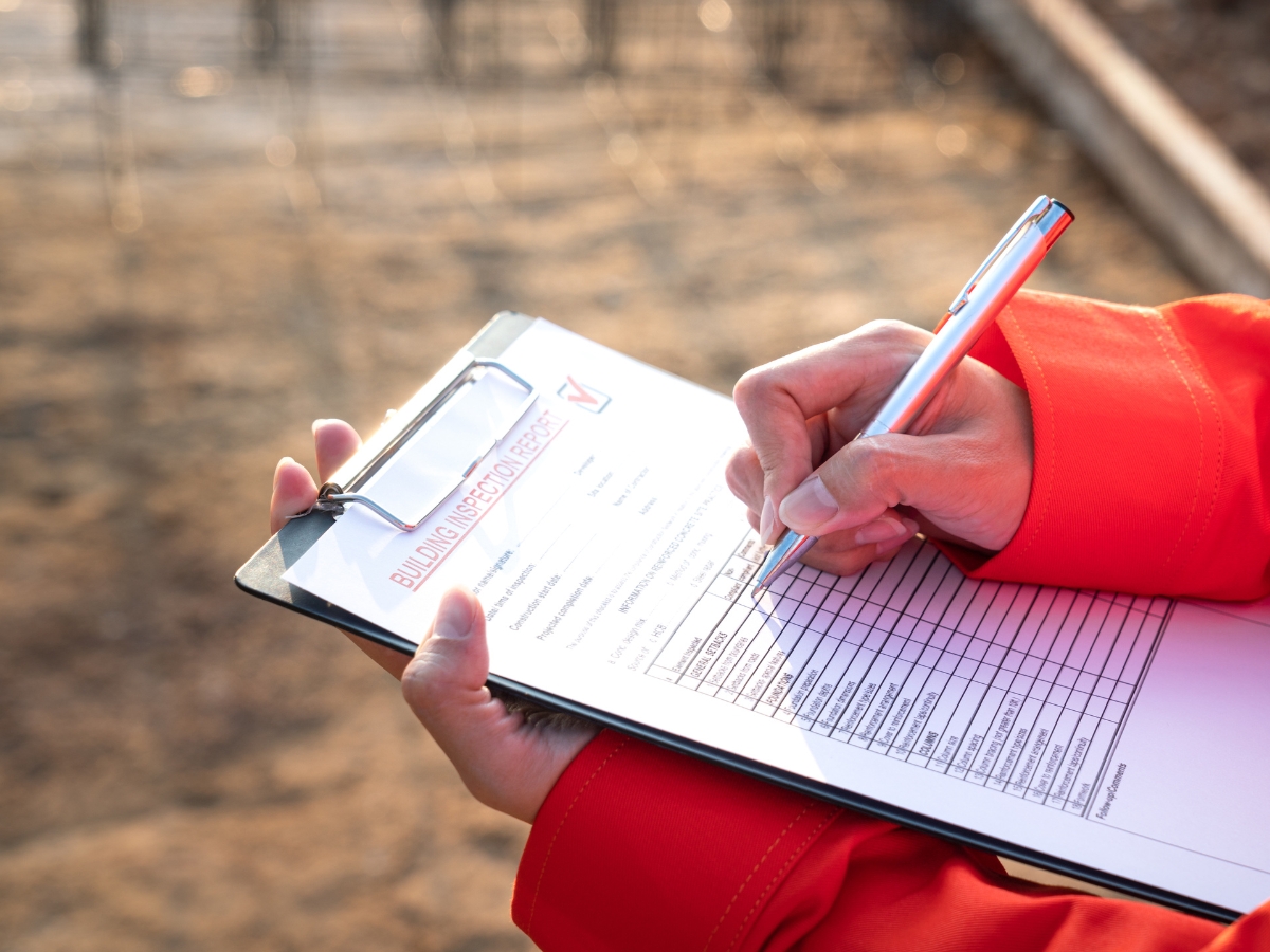 Close-up of a person in a bright orange jacket holding a clipboard, meticulously filling out a building inspection checklist with a pen. The background reveals the bustle of a construction site, emphasizing the thoroughness required for tasks like Professional Septic Inspection and Repair.