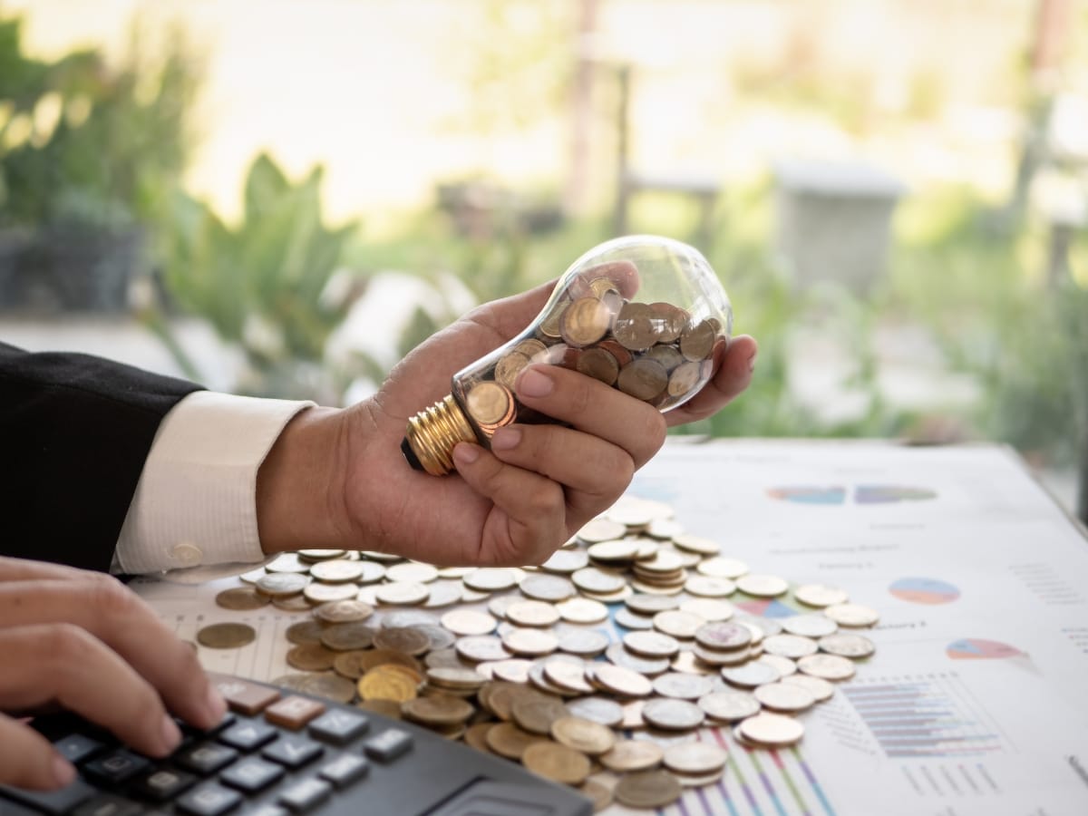 A hand holds a light bulb filled with coins, while another hand types on a calculator. More coins and financial documents are spread out on a table, emphasizing the importance of regular maintenance in managing finances effectively.