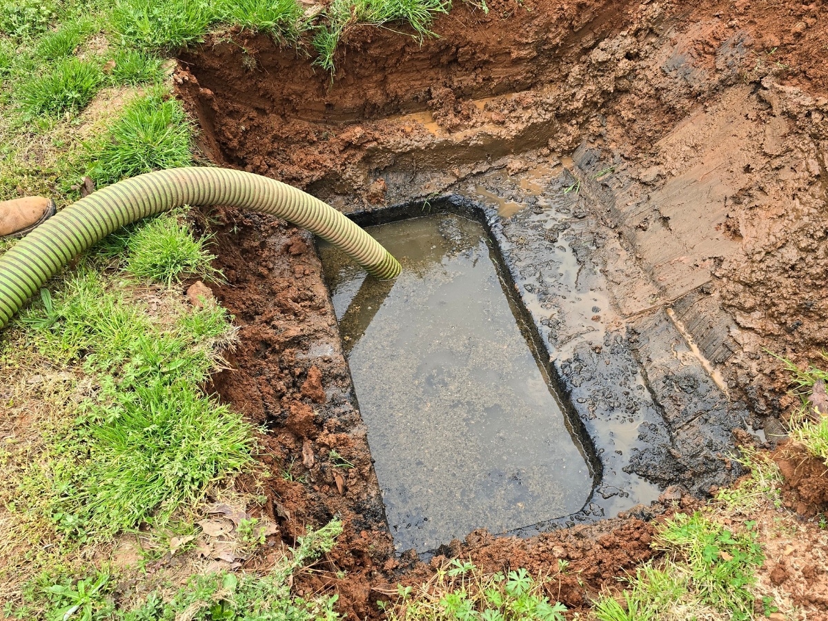 A rectangular hole in the ground filled with murky water, featuring a green hose inserted into it, surrounded by grass and dirt, highlights the importance of regular maintenance to avoid high septic system costs.