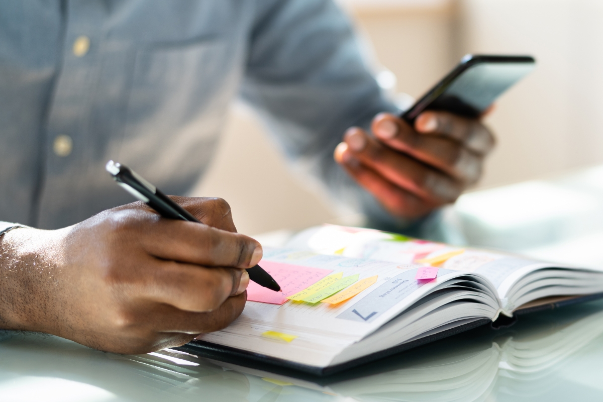 A person writes in an open notebook with one hand while holding a smartphone in the other, potentially searching for septic tank pumping frequency guidelines, surrounded by sticky notes.