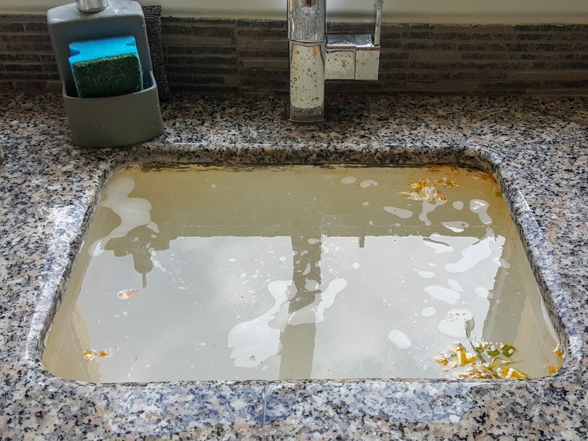 A kitchen sink filled with dirty water, food particles, and bubbles sits on a granite countertop, hinting at an urgent need for emergency septic tank cleaning; nearby, a sponge and soap dispenser stand ready.