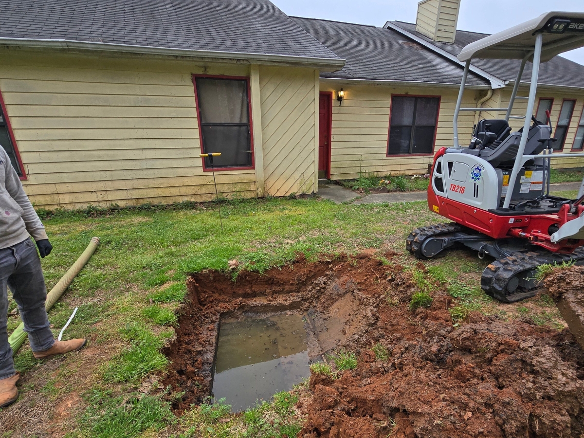 A hole filled with water is being excavated in the yard of a yellow house using a small excavator for a professional septic inspection and repair. A person is standing nearby, wearing gloves. The house has red-framed windows and a red door.