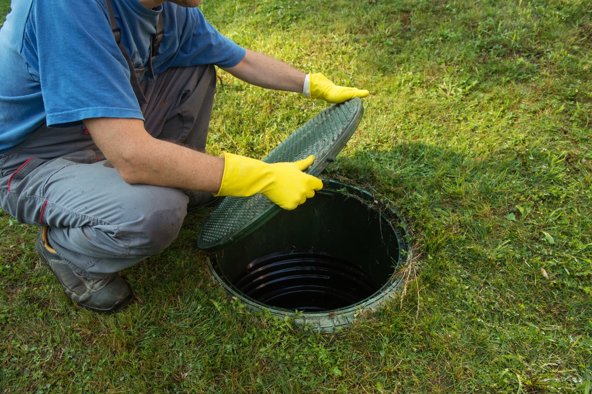 Person wearing yellow gloves and blue shirt lifting the lid of an outdoor septic tank on a grassy area, engaging in septic system troubleshooting.