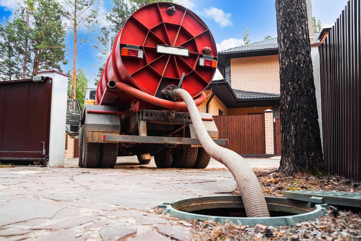 A large red septic truck is parked near a residential area, with a hose extending from the truck into an open sewer or septic tank on the ground as part of a septic system troubleshooting procedure.