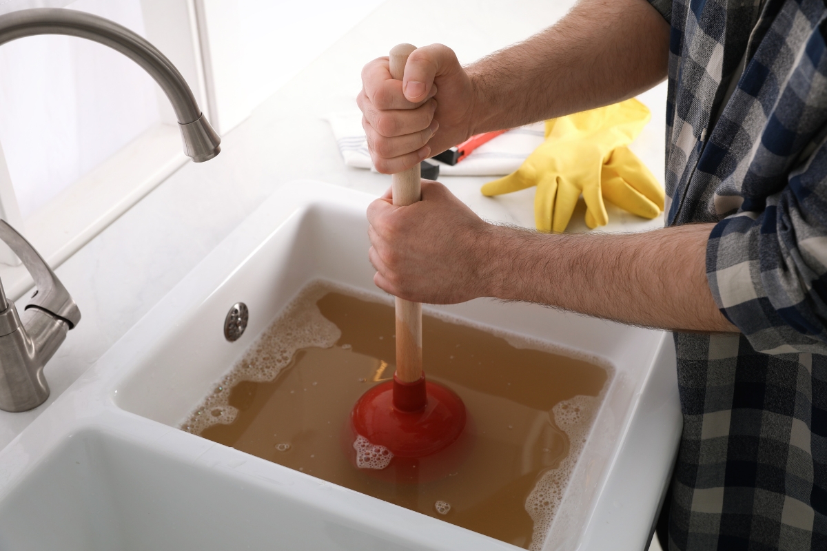 A person wearing a plaid shirt uses a plunger to clear a clogged kitchen sink filled with dirty water, showcasing their septic system troubleshooting skills, with yellow gloves resting on the counter beside the sink.