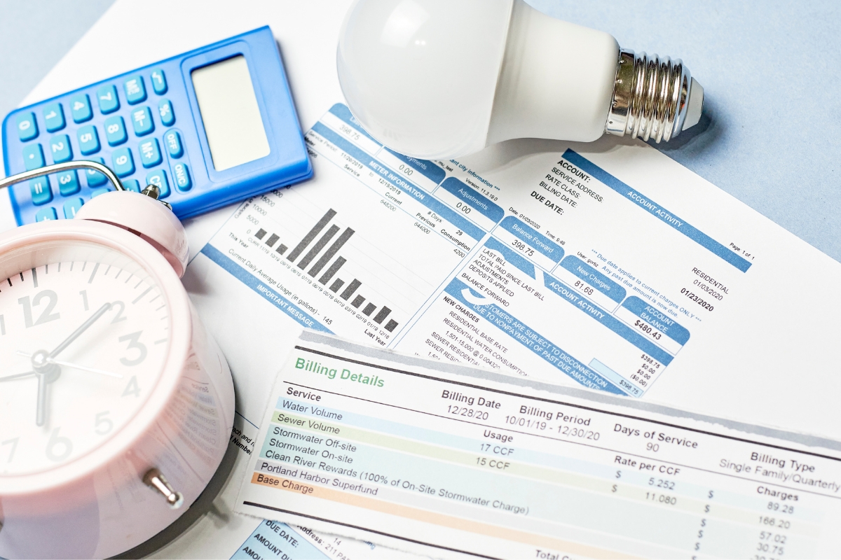 A clock, a calculator, a lightbulb, utility bills with bar graphs and billing details, and notes on septic system troubleshooting are scattered on the desk.