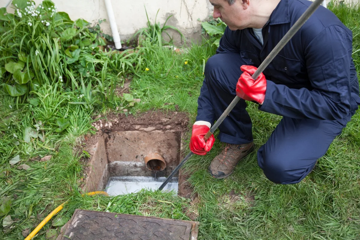 Septic System Troubleshooting: Expert Tips for a Smooth-Running System 6 A person wearing red gloves and blue overalls is using a long tool to clean or inspect a drain in a garden area, possibly involved in septic system troubleshooting.